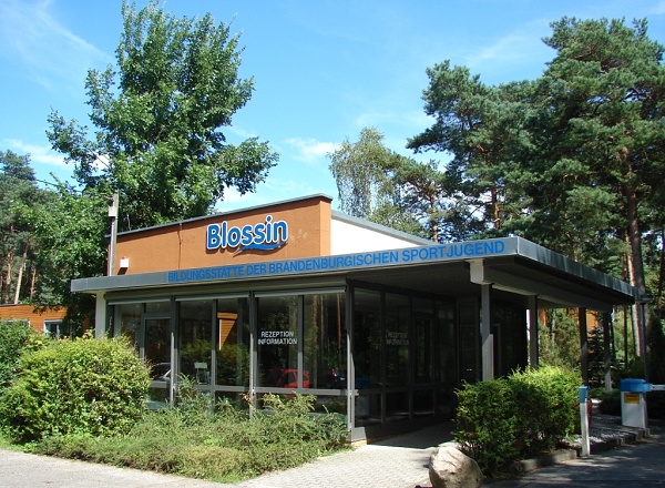 Eingangsbereich des Jugendbildungszentrums Blossin mit einem modernen Gebäude, großen Fenstern und einer überdachten Eingangstür, umgeben von Bäumen und einer begrünten Außenanlage