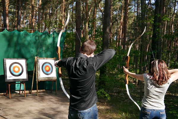 Zwei Personen schießen mit Pfeil und Bogen auf Zielscheiben im Wald.