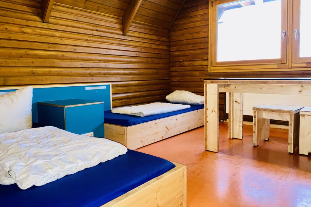 Untere Etage von einem Holzhaus im Jugenddorf. Zwei Betten und ein großer Tisch mit Hockern.