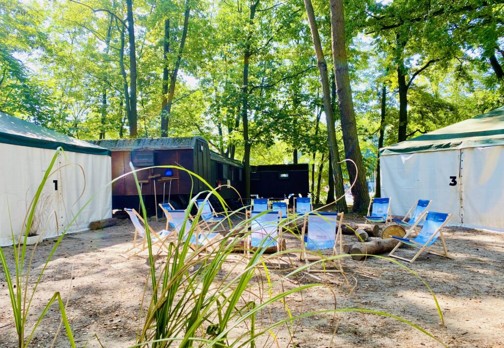 Zelte um einen Feuerplatz mit Sitzmöglichkeiten und Zirkuswagen im Hintegrund.